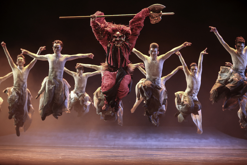 A group of dancers jump in the air. The central figure wheres a red costume and mask and holds a stick over their head.